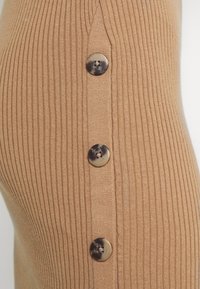 Vêtement en maille marron côtelée avec une coupe ajustée, comportant trois boutons ronds foncés sur le côté pour le détail. La texture est douce et extensible.