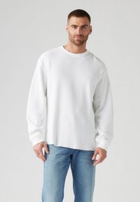 Hvit ribbet langermet skjorte med rund hals, kombinert med lyseblå denimjeans. Stoffet ser teksturert ut og har en litt løs passform.