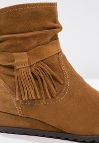 Bruine suède enkellaars met een gevouwen bovenkant, decoratieve franjes en stiksels. De zool is zwart en heeft een gestructureerd oppervlak voor grip.
