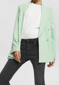 Femme portant un blazer croisé vert menthe, une chemise blanche et un jean noir taille haute avec braguette boutons sur un fond uni.