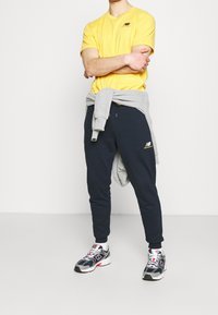 Geel t-shirt met korte mouwen, marineblauwe joggers, met een grijze sweatshirt om de taille geknoopt. Sneakers met een mix van grijze, marineblauwe en rode accenten.