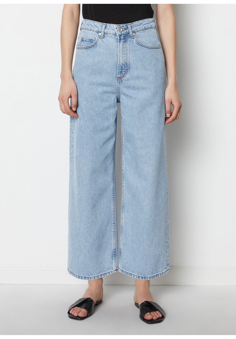 Femme portant un jean bleu clair à jambes larges et des sandales plates noires ouvertes à l'avant, debout contre un mur blanc uni.