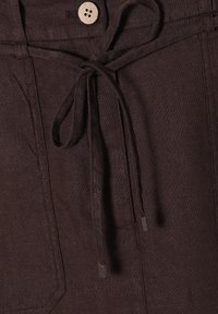 Smeđi laneni hlače s elastičnim pojasom, s drvenim dugmetom i teksturiranim materijalom. Uključuju bočne džepove i elegantne detalje.