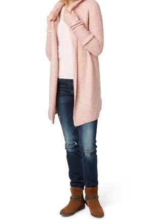 Personne portant un cardigan rose duveteux ouvert, un haut en maille blanc, un jean bleu foncé déchiré et des bottines marron, debout les mains tenant les bords du cardigan.