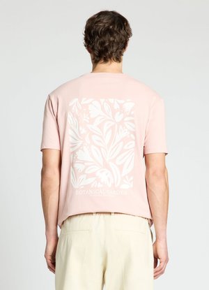 Mann trägt ein hellrosa Kurzarmhemd mit weißem botanischem Muster und Text, kombiniert mit hellbeiger Hose, Rücken zur Kamera.