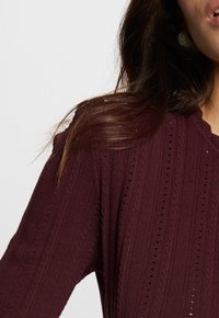 Nahaufnahme einer Person, die einen strukturierten, dunkelburgunderfarbenen Strickpullover trägt. Man sieht die Schulter, einen Teil des Gesichts und die langen dunklen Haare vor einem hellen Hintergrund.