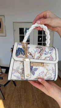 Sac à main blanc avec poignée tressée, imprimé floral, chaîne dorée et étiquette de marque Aldo, tenu par deux mains à l'intérieur.