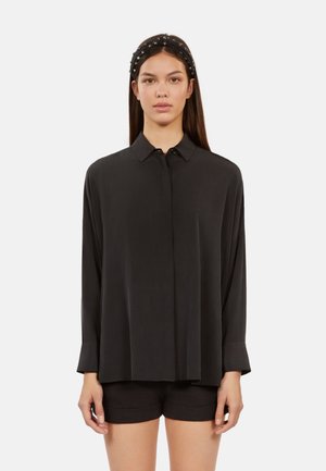 Chemise noire à manches longues avec un devant à boutons, en tissu doux, coupe décontractée et un détail de pli subtil. Portée avec un short noir et un bandeau.