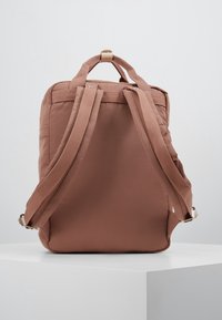 Ruskea kangasreppu, jossa on pehmustetut hihnat, yläkahva ja sileä rakenne. Ominaisuuksiin kuuluu yksinkertainen muotoilu ilman koristeellisia elementtejä.