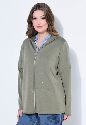 Vrouw draagt een olijfgroene hoodie met rits en kleine stippen over een lichtblauw shirt en blauwe bloemenbroek, staand tegen een witte achtergrond.
