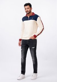 Sweatshirt crème à col zip quart avec des accents bleu marine et bordeaux. Porté avec un jean noir déchiré et des baskets blanches. Logo doré sur la poitrine.