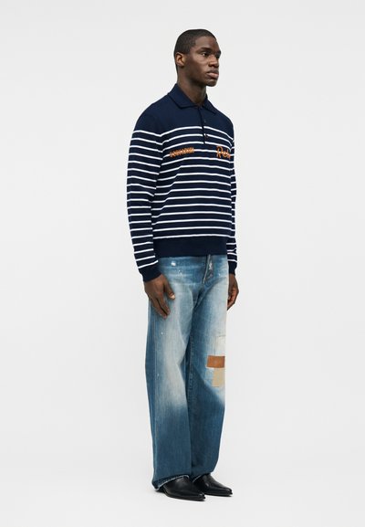 Homme portant un polo à manches longues rayé bleu marine et un jean ample déchiré avec des patchs, debout devant un fond blanc uni.