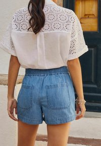 Valkoinen paita, jossa on rei'ityksellä laittuja yksityiskohtia ja lyhyet hihat, yhdistettynä sinisiin farkkushortsihin, joissa on joustava vyötärö ja kaksi takataskua.