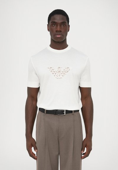 Hvid kortærmet t-shirt med et tekstureret logo design på brystet, parret med lysebrune skræddersyede bukser og et sort bælte.