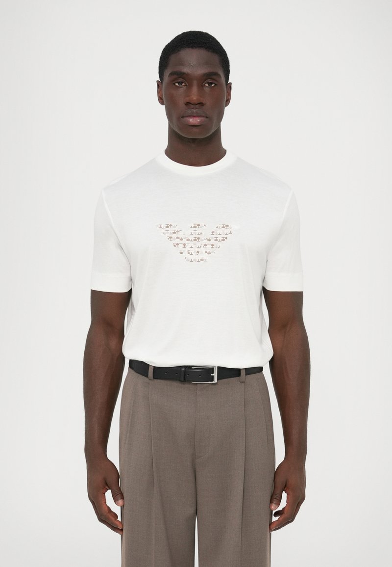 Λευκό κοντομάνικο t-shirt με ανάγλυφο σχέδιο λογότυπου στο στήθος, συνδυασμένο με ανοιχτό καφέ προσαρμοσμένα παντελόνια και μαύρη ζώνη.
