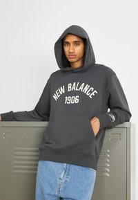 Sudadera gris oscura con un bolsillo frontal y "NEW BALANCE 1906" en texto blanco. Presenta un corte holgado y puños acanalados. Llevada sobre jeans azules.