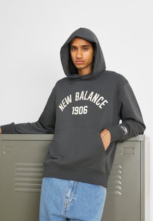 Sudadera gris oscura con un bolsillo frontal y "NEW BALANCE 1906" en texto blanco. Presenta un corte holgado y puños acanalados. Llevada sobre jeans azules.