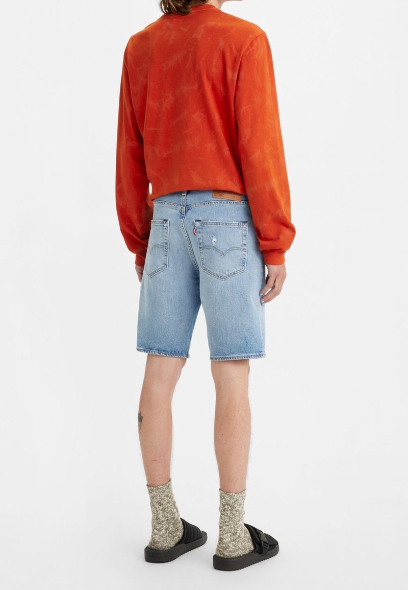 Persona con camisa de manga larga naranja, pantalones cortos de mezclilla azul claro, calcetas estampadas y sandalias negras, de pie mirando hacia atrás sobre un fondo blanco.