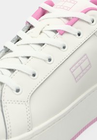 Valkoinen tenispuku vaaleanpunaisilla yksityiskohdilla, valkoiset nauhat ja "Tommy Jeans" -logo sivulla, esitetty lähikuvassa kulma-näkymässä.
