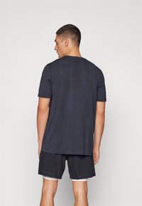 Sötétkék, rövid ujjú póló, texturált anyagból, a hátsó részén függőleges "UNDER ARMOUR" logóval, fekete rövidnadrággal párosítva.