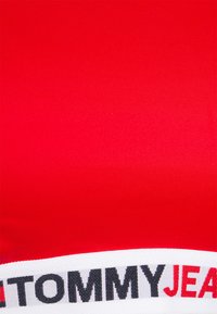 Sutiã desportivo de tecido elástico vermelho com uma faixa elástica branca apresentando "TOMMYJEANS" em letras pretas e vermelhas na parte inferior.