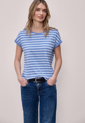 Młoda kobieta o blond włosach, ubrana w niebiesko-białą, paskowaną koszulkę wpuszczoną w niebieskie dżinsy, z rękami w kieszeniach, stojąca na jasnym tle.