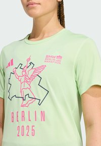 Vaaleanvihreä urheilupaita, jossa on graafinen kuvio pinkistä enkeli-hahmosta, teksti "BMW BERLIN MARATHON" ja "2025."