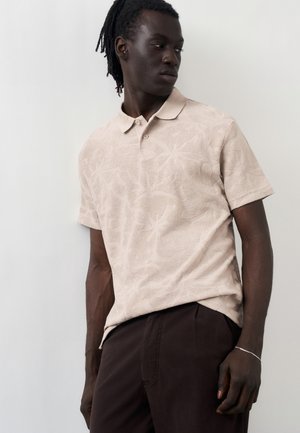 Άνδρας με σκουρόχρωμο δέρμα και dreadlocks, φορώντας μια μπεζ κοντομάνικη πόλο με υφή λουλουδιών και σκούρο καφέ παντελόνι, στέκεται μπροστά σε έναν λευκό τοίχο.