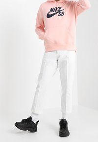 Person som bär en rosa Nike SB-huvtröja, vita korta byxor, vita strumpor och svarta sneakers står mot en vit bakgrund.