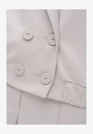 Beżowa marynarka dwurzędowa z czterema okrągłymi, żebrowanymi guzikami oraz dopasowane, skrojone na miarę spodnie w zbliżeniu.