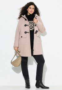 Abrigo duffle de color rosa claro con botones negros, dos bolsillos frontales, capucha, usado sobre un suéter negro con estampado y jeans oscuros, sosteniendo una bolsa beige.