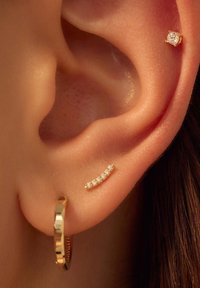 Gold hoop earring with a textured band and a small curved stud featuring five sparkling clear stones, set against a close-up of ears.