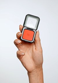 Vierkante zwarte compact met een transparante deksel, die een levendige oranje blush bevat. De hand die het vasthoudt heeft lichtgekleurde, verzorgde nagels.