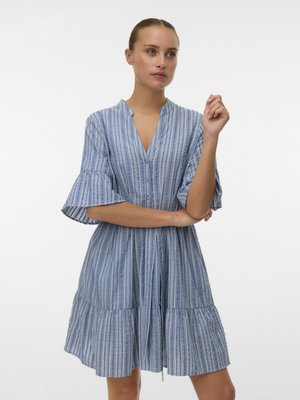 Kobieta z włosami związanymi w kok, ubrana w niebiesko-białą sukienkę w pionowe paski z rozkloszowanymi rękawami i warstwową spódnicą, stojąca z założonymi ramionami.