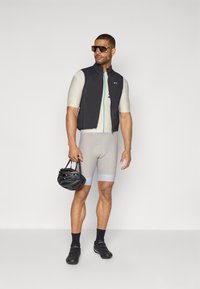 Gilet de cyclisme noir avec fermeture éclair à l'avant, maillot ajusté beige clair, shorts gris et chaussures de cyclisme noires. Tient un casque noir.