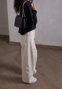 Haut noir à manches longues, pantalon beige ample, baskets blanches et un sac à main noir matelassé avec une bandoulière en chaîne dorée. Fond en parquet.