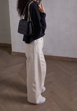 Haut noir à manches longues, pantalon beige ample, baskets blanches et un sac à main noir matelassé avec une bandoulière en chaîne dorée. Fond en parquet.