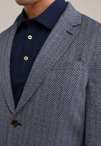 Marineblauwe herringbone jas met een gestructureerd patroon, enkele knoopsluiting en notch revers, gecombineerd met een donkerblauwe overhemd met kraag.