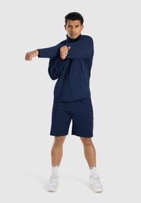 Chaqueta atlética azul marino con cuello alto, bolsillos con cremallera y pantalones cortos a juego en azul marino. El modelo realiza una pose de estiramiento de brazo.