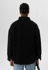 Zwarte oversized shirt met een kraag, met een zachte textuur, contrasterende stiksels en zijsplitten bij de mouwen. Draagt met lichtblauwe jeans.