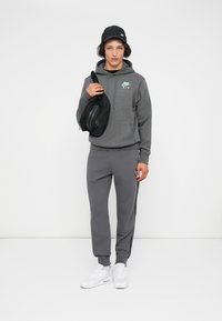Sweater cinza com logótipo, calças de treino cinzas, ténis brancos e uma mala cruzada preta. Modelo a usar um boné preto, de pé contra um fundo simples.