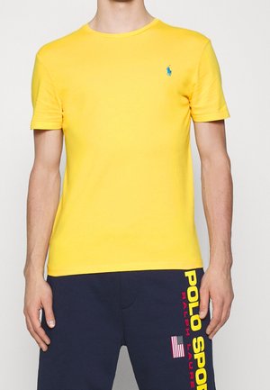 Homme portant un t-shirt jaune à manches courtes avec un petit logo bleu, associé à un short bleu marine avec un texte "Polo Sport" jaune et rouge en gras, ainsi qu'un drapeau américain.