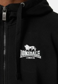 Sort zip-up hoodie lavet af blødt stof, med et hvidt broderet løve-logo og teksten "Lonsdale London" på brystet. Sølv lynlåsdetalje inkluderet.