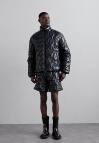 Schwarze gestepptes Pufferjacke mit Zick-Zack-Muster, kombiniert mit passenden Shorts und schwarzen wasserdichten Stiefeln. Ausgestattet mit einem hohen Kragen und einem Reißverschluss.
