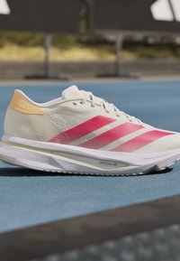 Λευκό και ροζ παπούτσι για τρέξιμο Adidas με σόλα Lightstrike Pro σε μπλε επιφάνεια τροχιάς, λεπτομερής πλευρική όψη.