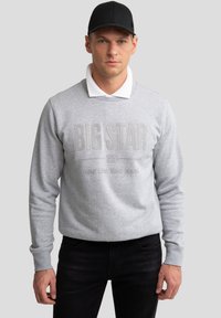 Mand iført sort kasket, grå sweatshirt med teksten "BIG STAR", hvid skjortekrave og sorte bukser, stående foran en grå baggrund.