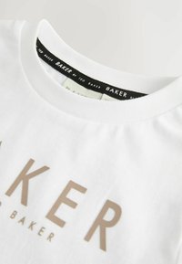 Valge puuvillane T-särk, millel on ümar kaelus, must silt ja ees suur beež "BAKER" tekst. Sile tekstuur, minimaalne disain.