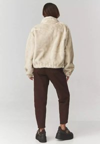 Kremowa kurtka z sztucznego futra z wysokim kołnierzem, elastycznym dołem i luźnym krojem, zestawiona z ciemnobrązowymi zwężanymi spodniami oraz masywnymi czarnymi sneakersami.