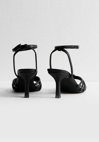 Czarne sandały na wysokim obcasie z paskowym wzorem, regulowanym paskiem na kostce i zwężanym obcasem. Gładka skórzana faktura i minimalne detale.
