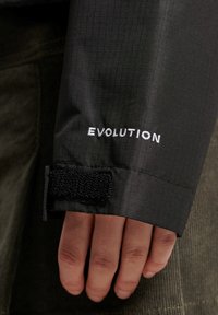 Zwarte stoffen jacksleeve met een gestructureerd ruitpatroon, voorzien van een witte geborduurde "EVOLUTION" logo en een zwarte klittenbandsluiting.